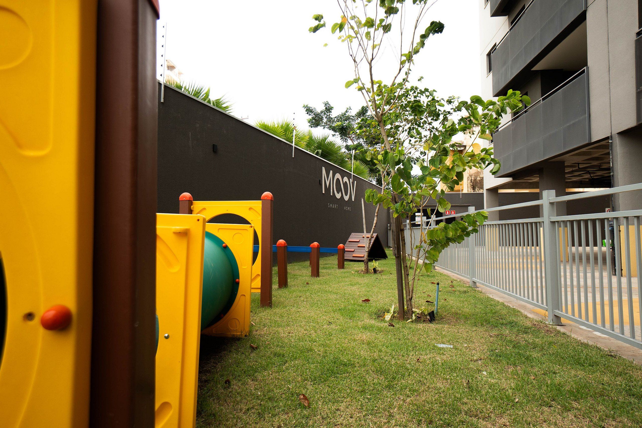 Un parque infantil con estructuras de juego amarillas y un árbol pequeño en un espacio cubierto de césped.