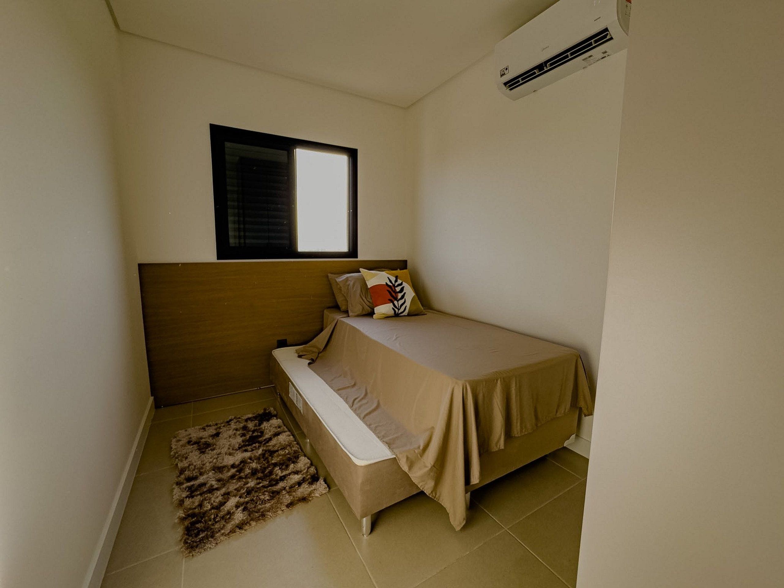 A small, single-bed room with a window, a wall-mounted air conditioner, and a rug on the floor.
