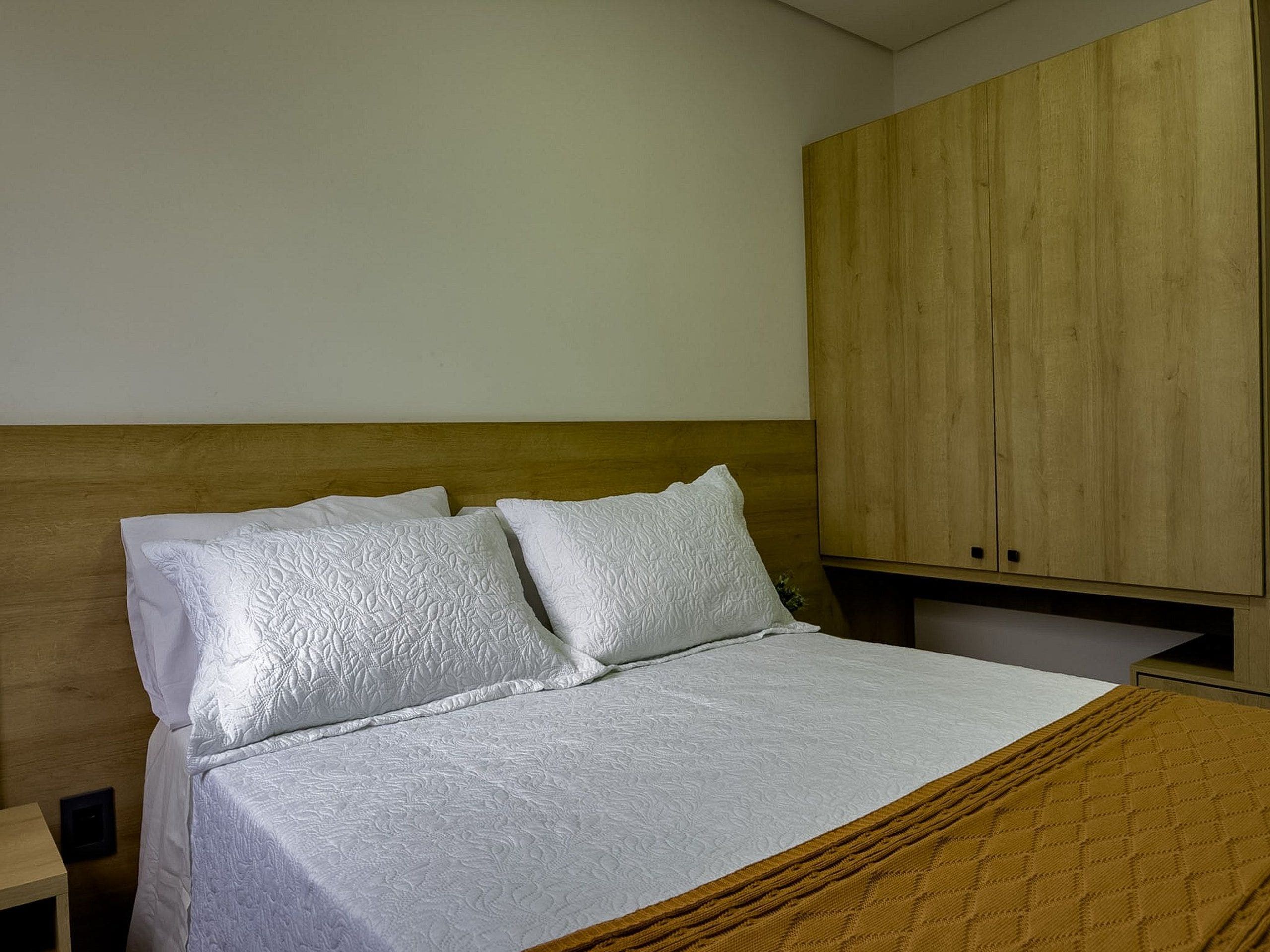 Una cama con colcha blanca acolchada y dos almohadas, cabecera de madera y un armario de madera en una habitación con paredes lisas.