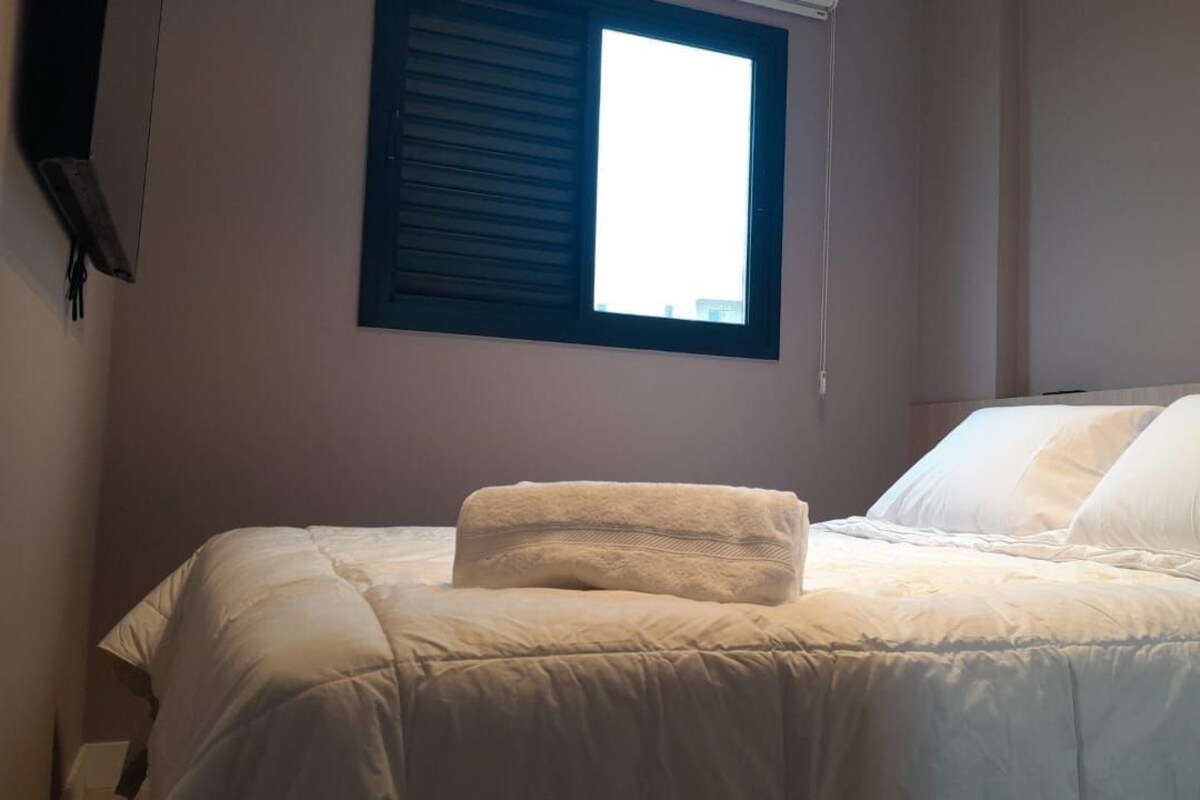 A bed with a white comforter and pillows, a window with closed blinds, and a wall-mounted light fixture.