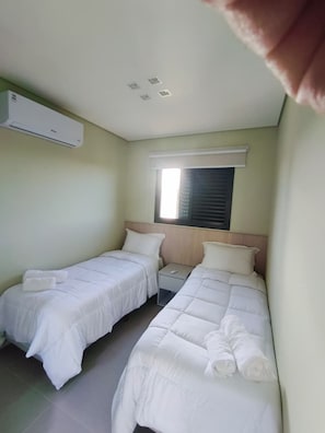 Habitación de hotel con dos camas, una ventana con persianas, una mesita de noche y un aire acondicionado de pared.