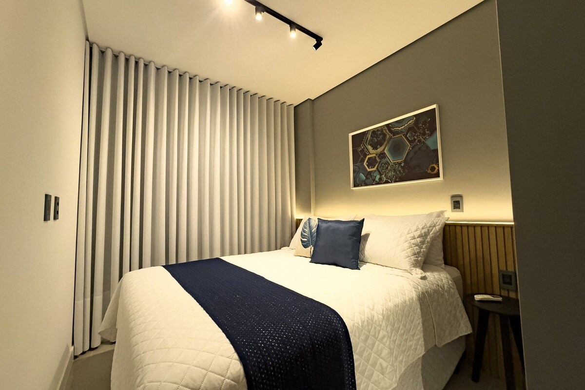 A hotel room with a large bed, a bedside table, a wall-mounted artwork, and a window with blinds.