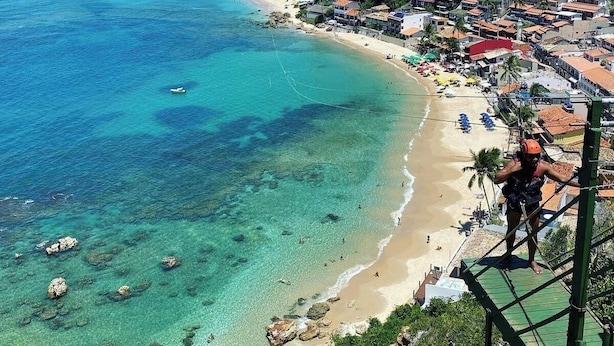 Un pueblo costero con playa, aguas cristalinas y una persona en una tirolesa.