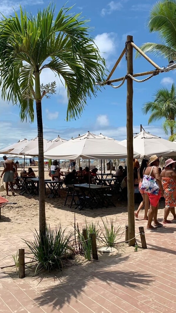 A beachside dining area with tables and umbrellas, surrounded by palm trees.