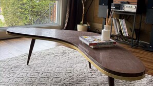 Ein Zimmer mit einem gebogenen Holztisch, einer Topfpflanze, einer kleinen weißen Vase und einem Bücherregal mit Büchern und einem Plattenspieler.