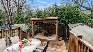 Eine Holzterrasse mit einem für eine Mahlzeit gedeckten Tisch, einem überdachten Liegestuhl und einer überdachten Sitzgruppe.