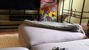 Ein Wohnzimmer mit einer Couch, einer Decke, einem Spielcontroller und einem Spider-Man-Comic auf einem Regal.