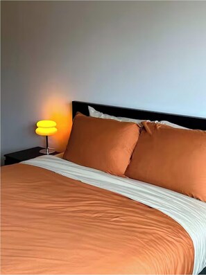Una cama con una colcha blanca y naranja, dos almohadas naranjas y una mesita de noche con una lámpara amarilla.