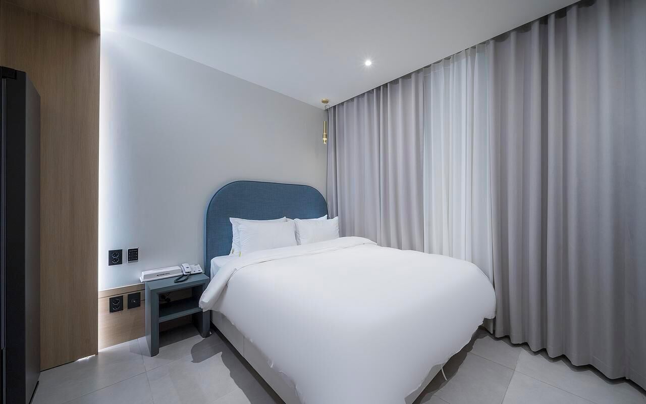 A modern hotel room with a large bed, a nightstand, a desk, and a window with curtains.