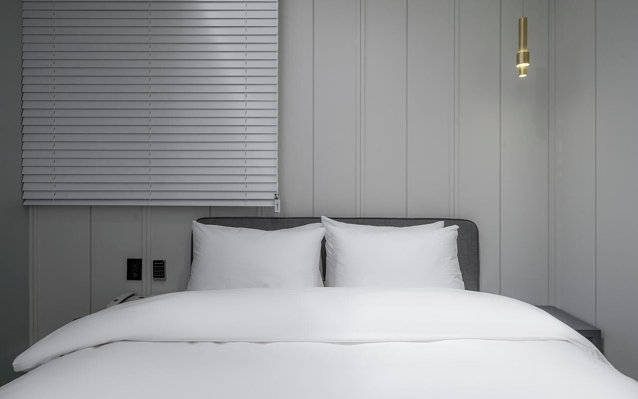 Habitación de hotel con cama, ropa de cama blanca, cabecero, lámpara de pared y ventana con persianas.