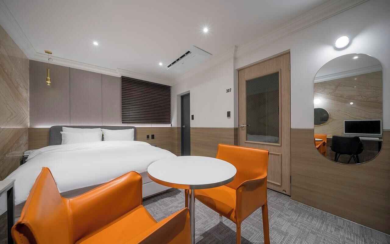 Habitación de hotel con una cama, dos sillas naranjas, una mesita redonda, un televisor y un espejo.