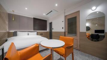 Habitación de hotel con una cama, dos sillas naranjas, una mesita redonda, un televisor y un espejo.