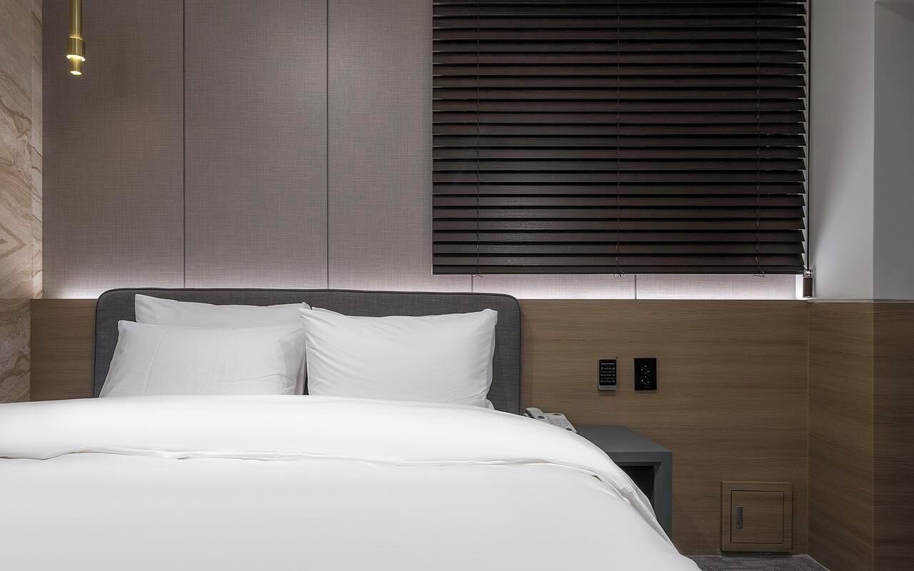 A modern hotel room with a large bed, a bedside table, a phone, and a wall-mounted control panel.
