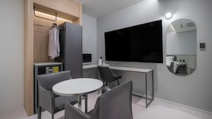 A modern hotel room with a large flat-screen TV, a round dining table, and a small kitchenette.