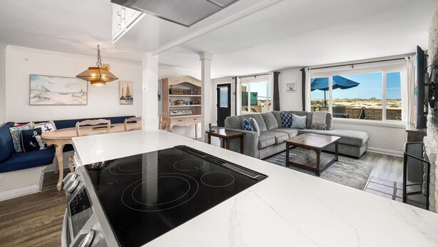 A modern kitchen with a central island, a dining area with a wooden table, a living room with a sofa and a coffee table, and large windows with a view of the beach. - Oceanfront Stay Near Newport Boardwalk (Newport Beach)