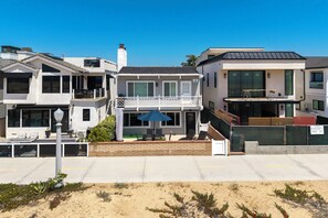 Ein zweistöckiges Haus mit einem Balkon, einer Terrasse mit Gartenmöbeln und einer sandigen Fläche im Vordergrund.