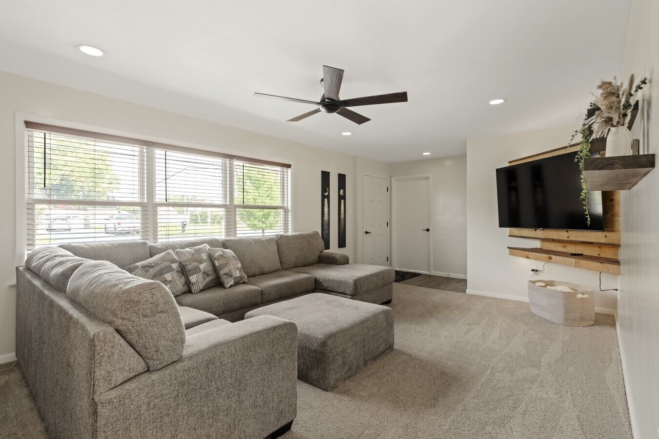 A modern living room with a sectional sofa, a ceiling fan, a flat-screen TV mounted on a wooden shelf, and a large window with blinds.