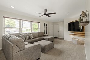 A modern living room with a sectional sofa, a ceiling fan, a flat-screen TV mounted on a wooden shelf, and a large window with blinds. - Cozy Family Getaway: Amish Farmland|Patio|Fire Pit|Central Lancaster County (Ronks)