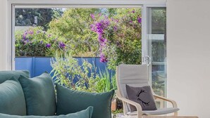 A room with a sofa, a chair, and a table. There is a glass door leading to an outdoor area with plants and flowers.