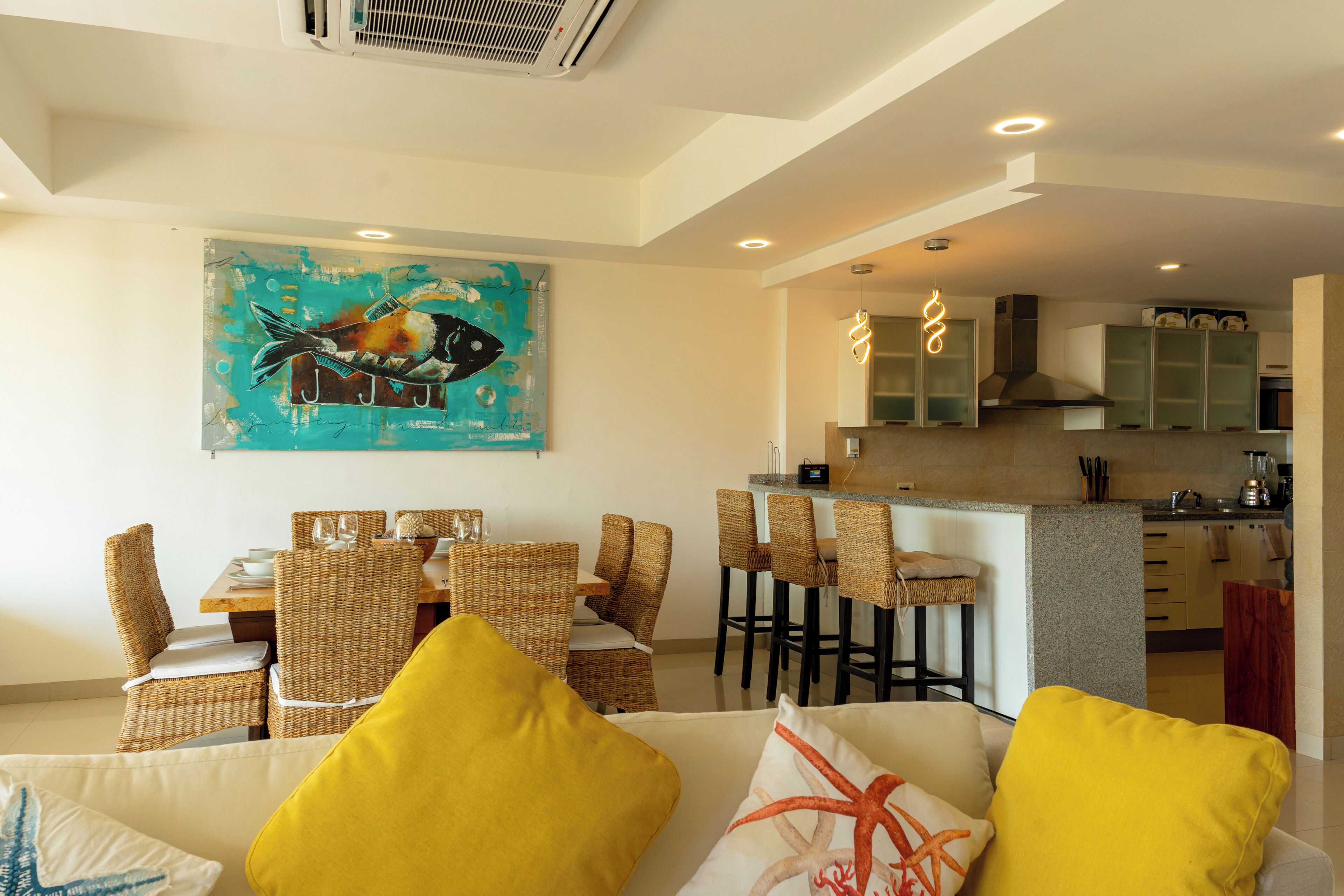 A dining area with wicker chairs and a table, a kitchen with a bar, and a living area with a sofa and cushions.