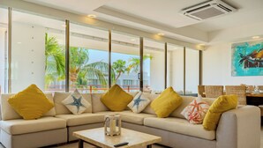 Una sala de estar moderna con sofá, cojines, una mesa de centro y vistas a palmeras a través de grandes ventanales.