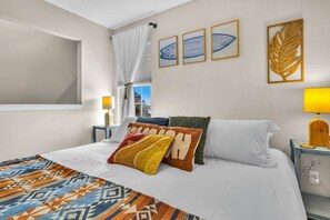 Ein Hotelzimmer mit Bett, Kissen, einer gemusterten Decke, einem Fenster mit Vorhängen, Wandbildern und einem Nachttisch mit Lampe.
