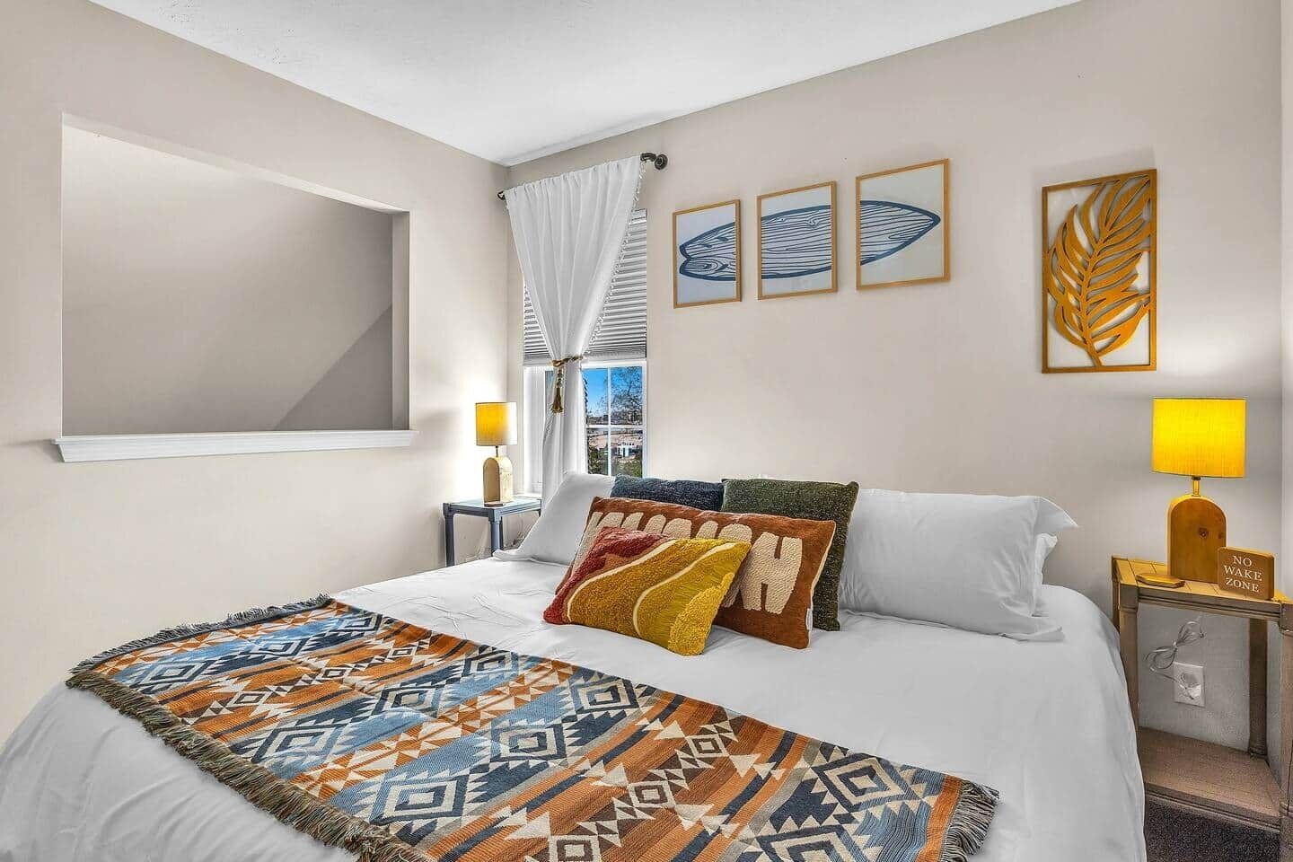 A hotel room with a bed, a window with curtains, a bedside table with a lamp, and wall art.