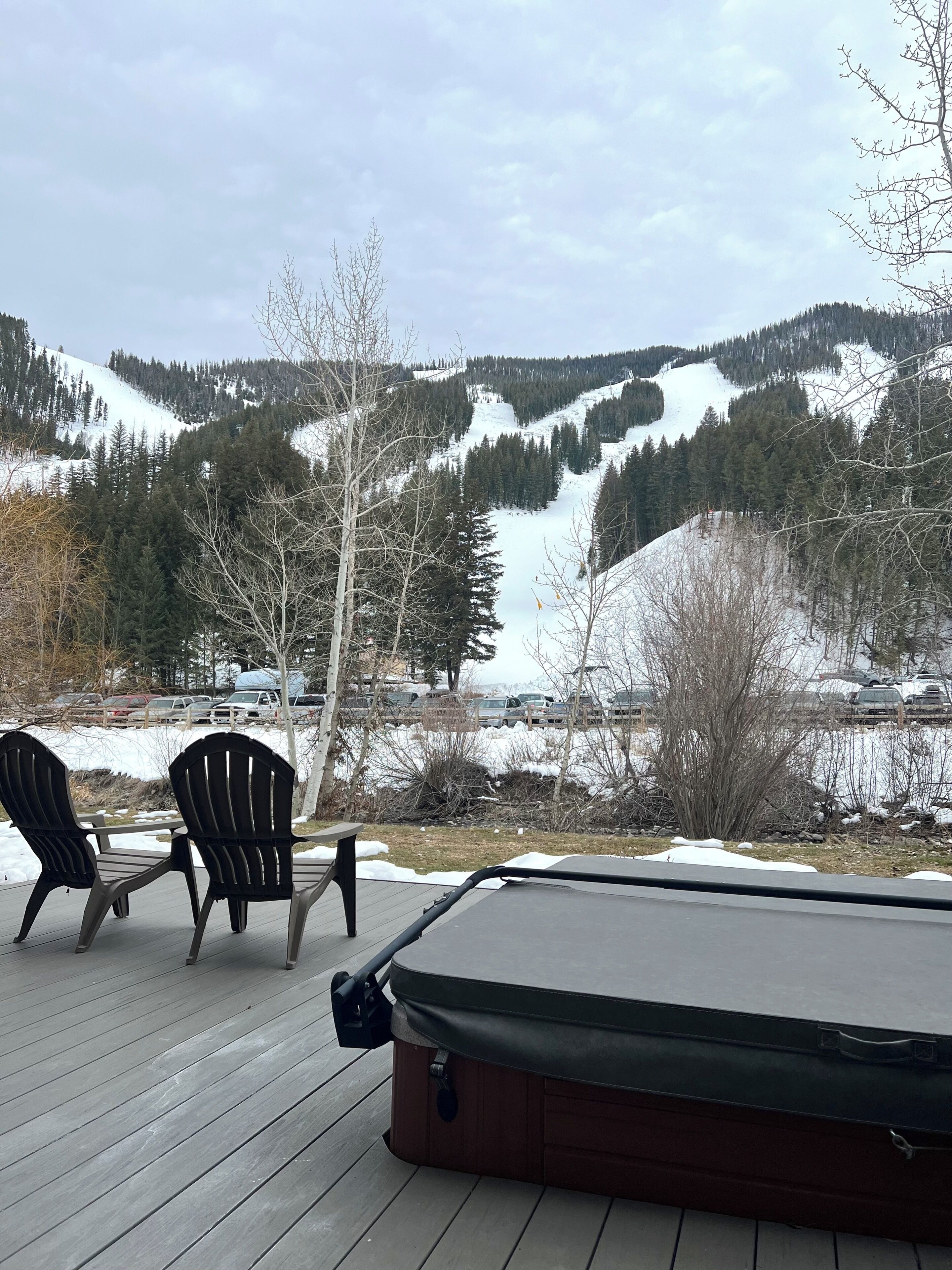 A deck with two Adirondack chairs and a hot tub overlooking a snowy mountain landscape.
