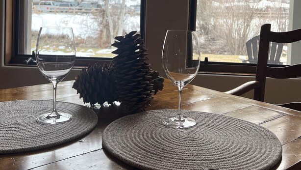 Une table à manger en bois, agrémentée de deux verres à vin et d’un centre de table en forme de pomme de pin, placée devant une fenêtre donnant sur un paysage enneigé.