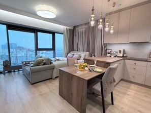 Una sala de estar moderna con un sofá, una mesa de comedor con sillas, una zona de cocina y ventanas amplias que ofrecen vistas de la ciudad.