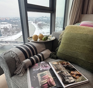 A window seat with a view of a snowy landscape, a plate with fruits, a magazine, and a green throw pillow. - Subtelna Elegancja Olszynki Park (Rzeszów)