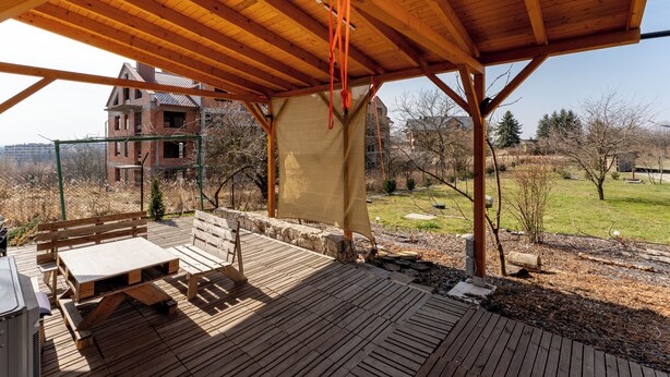 Eine überdachte Terrasse im Freien mit Holzmöbeln, Blick auf ein Haus im Hintergrund und eine Rasenfläche mit Bäumen.