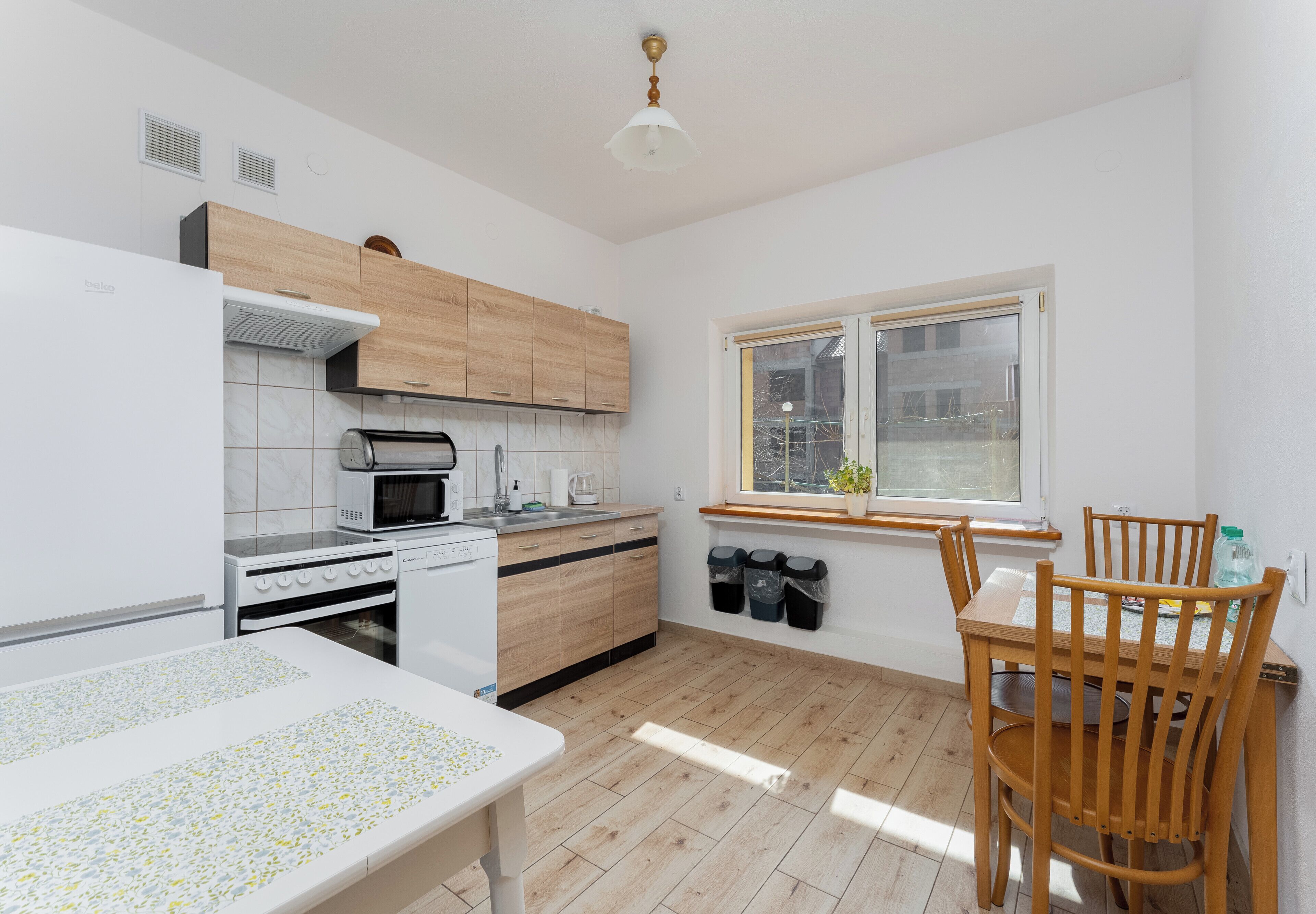Een moderne keuken met witte apparaten, houten kastjes, een eettafel met stoelen en een raam met jaloezieën.