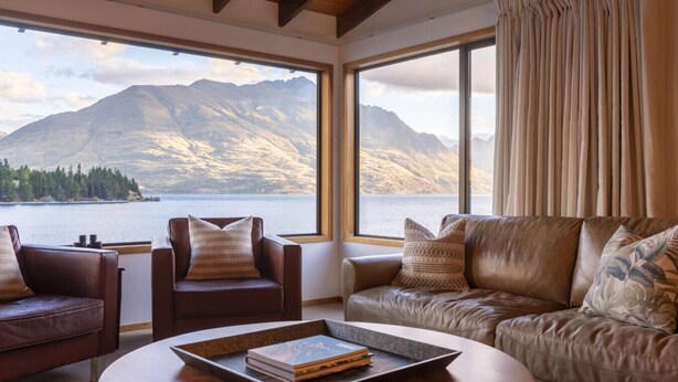 A room with a leather sofa, a leather armchair, and a coffee table with a book. Large windows offer a view of mountains and a lake. - The Art House | Queenstown lakefront & central location (Queenstown)