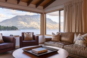 A room with a leather sofa, a leather armchair, and a coffee table with a book. Large windows offer a view of mountains and a lake. - The Art House | Queenstown lakefront & central location (Queenstown)