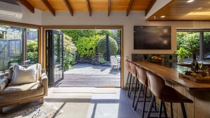 A modern kitchen with a bar area, a dining area with a sofa and chairs, and a view of a garden through glass doors. - The Art House | Queenstown lakefront & central location (Queenstown)