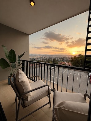 A balcony with wicker furniture, a potted plant, and a cityscape view at sunset.