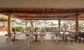 Un espace de restauration en plein air avec des tables et des chaises, un toit de chaume et des palmiers en arrière-plan.
