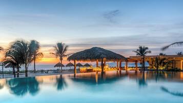 Área de piscina com vista para o pôr do sol, palmeiras e uma estrutura com telhado de palha.