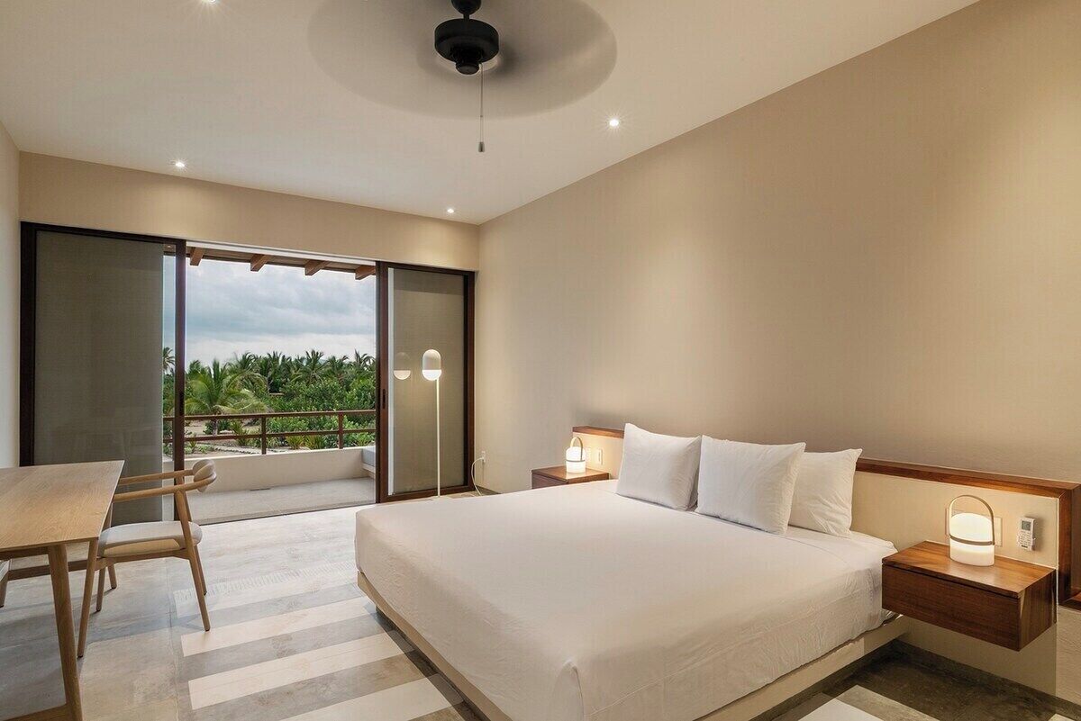 Quarto de hotel com cama, mesas de cabeceira, uma mesa com cadeiras e vista para o verde lá fora.