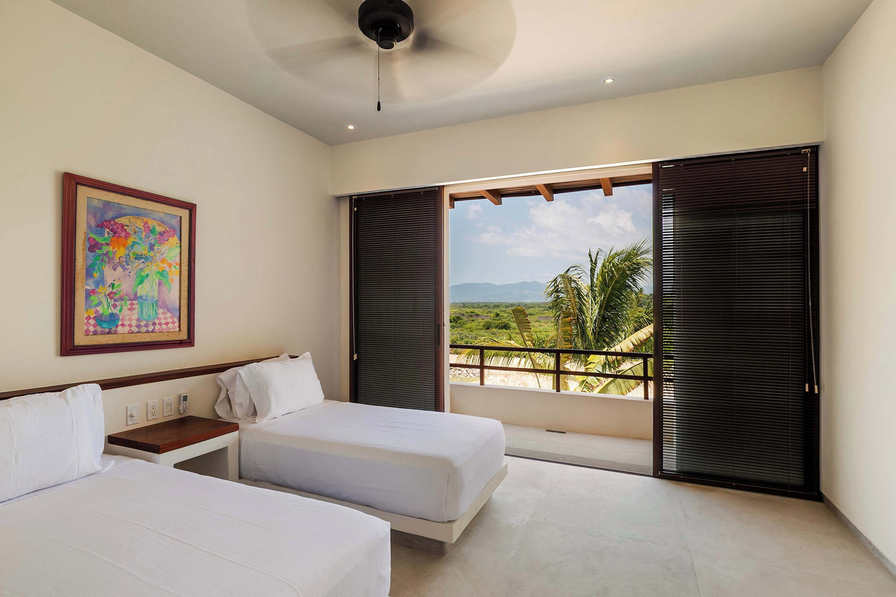 Une chambre d’hôtel avec deux lits, un ventilateur de plafond, un tableau et un balcon donnant sur le paysage.