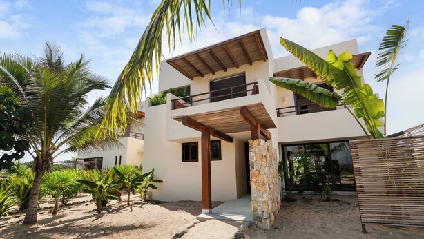 A modern two-story building with a balcony, surrounded by tropical vegetation and a sandy area.