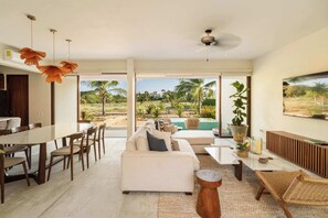 A modern living room with a sofa, dining table, and chairs, a ceiling fan, and a view of a tropical landscape. - Ground-Floor 4BR Ocean-View Villa | Marea (Zihuatanejo)