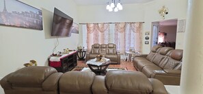 Ein Wohnzimmer mit braunen Ledersofas, einem Flachbildfernseher, einer Chandelüster und einem Essbereich mit Tisch und Stühlen.