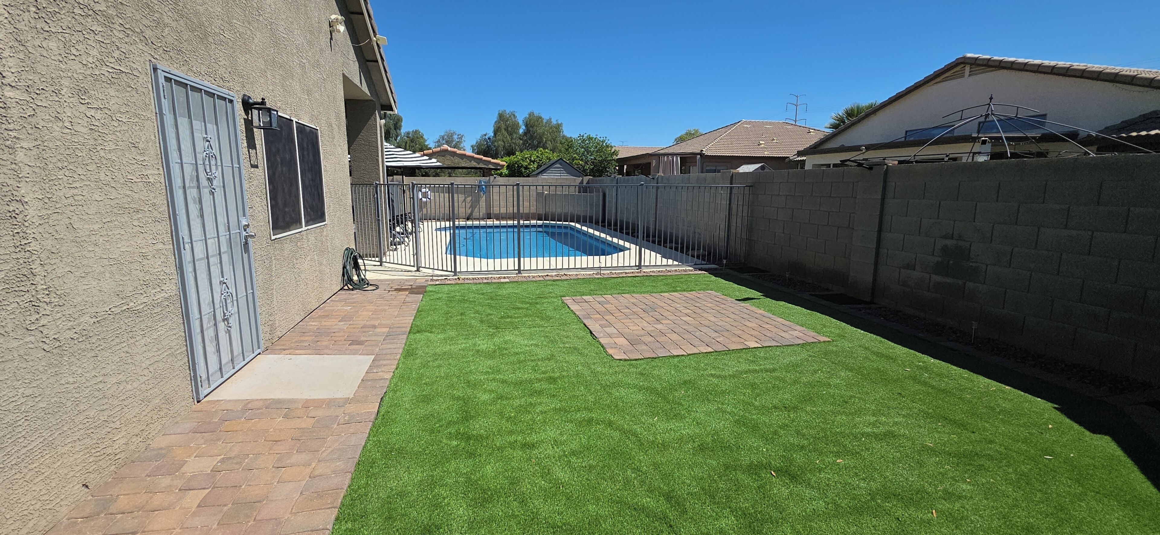 A backyard with a swimming pool, artificial grass, and a brick patio.