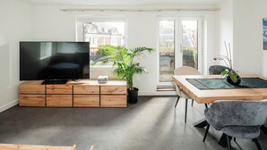 Ein modernes Wohnzimmer mit einem Flachbildfernseher auf einer Holzkommode, einem Esstisch mit Stühlen, einer Topfpflanze und Blick auf Gebäude durch ein großes Fenster.