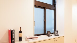 Um quarto com janela, uma garrafa de vinho, livros e quadros em um balcão.
