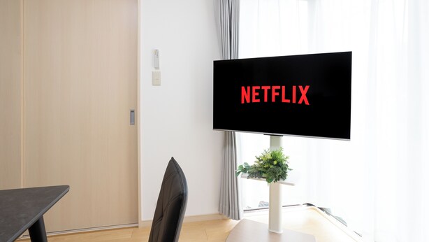 Quarto com televisão exibindo 'NETFLIX', uma escrivaninha, uma cadeira, uma planta e uma janela com cortinas.