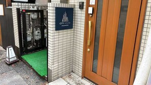 Entrada de um hotel com porta de madeira, portão de metal e uma placa com o texto 'CITY HOUSE KAGURAZAKA'.