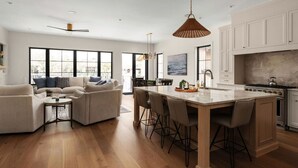 A modern kitchen with a central island, wooden cabinets, and a dining area with a sofa and chairs. - Luxury 3-Story Lakehouse |Pool + Hot Tub + Sundeck (Horseshoe Bay)
